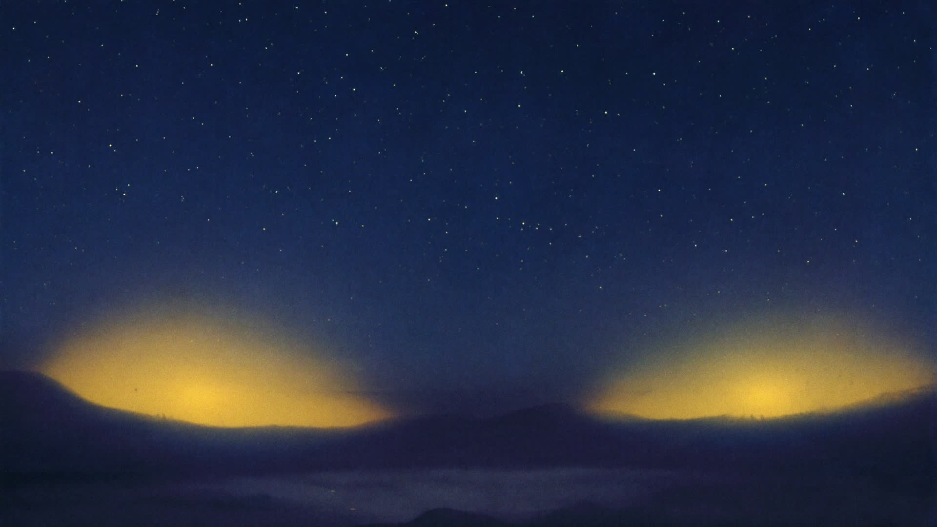 Two hills at night, each with a golden glow behind it, under a wide, empty, starry sky